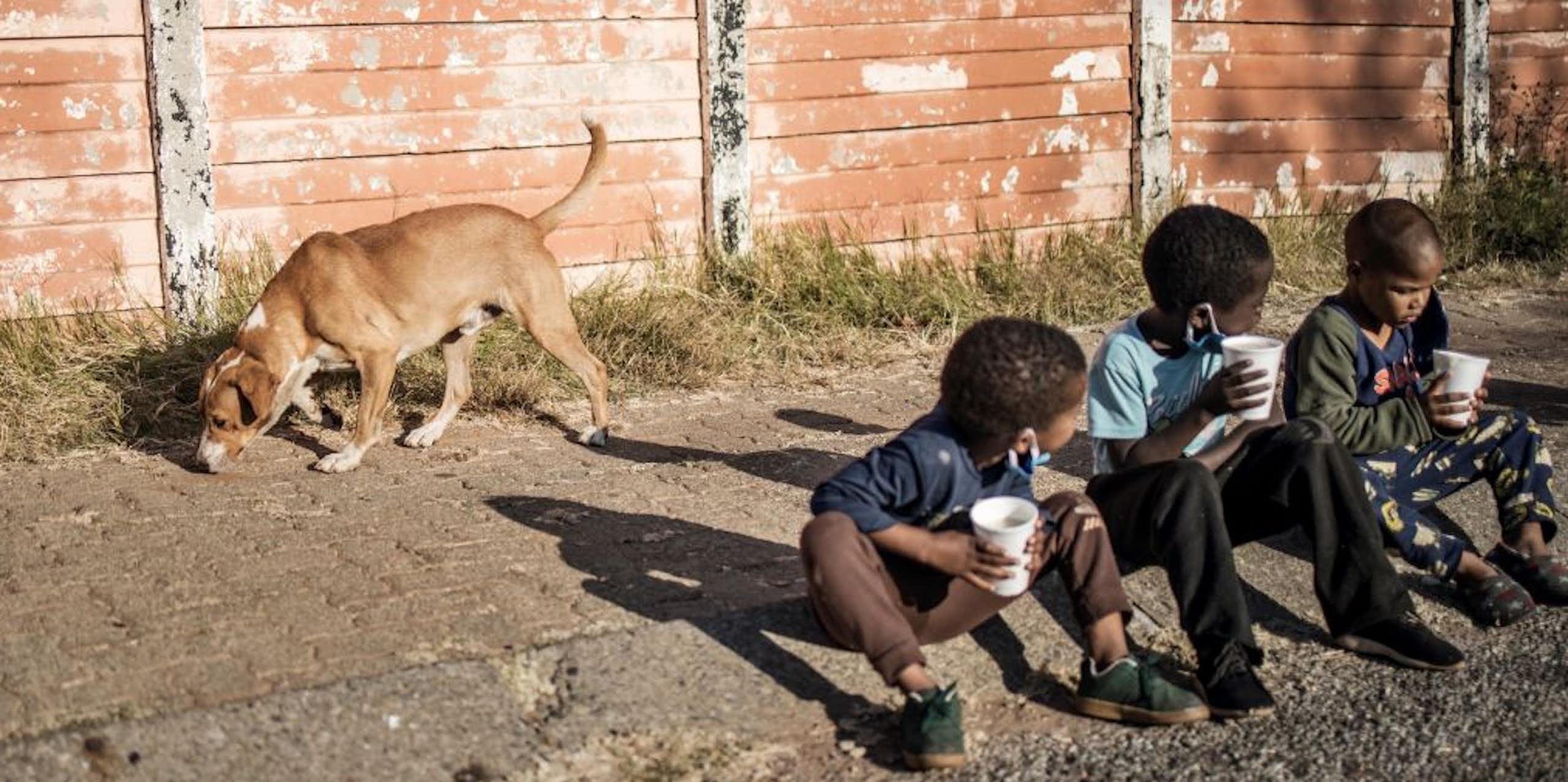 Three young children sit on a pavement, holding disposable cups, while a dog sniffs the ground behind them