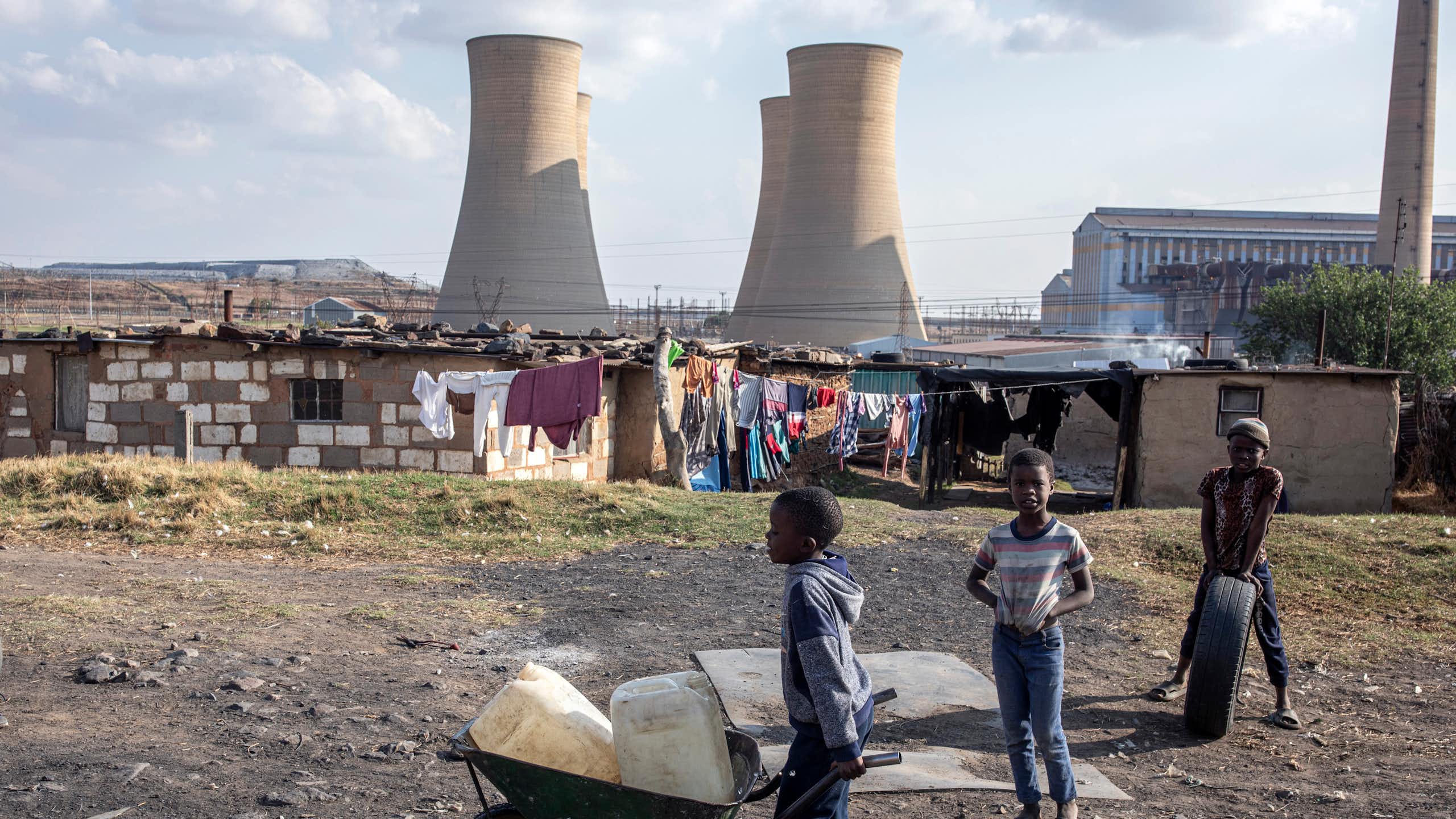 A coal fired power station with enormous towers and tin shacks built in front. A small boy wheels containers of water in a wheelbarrow.