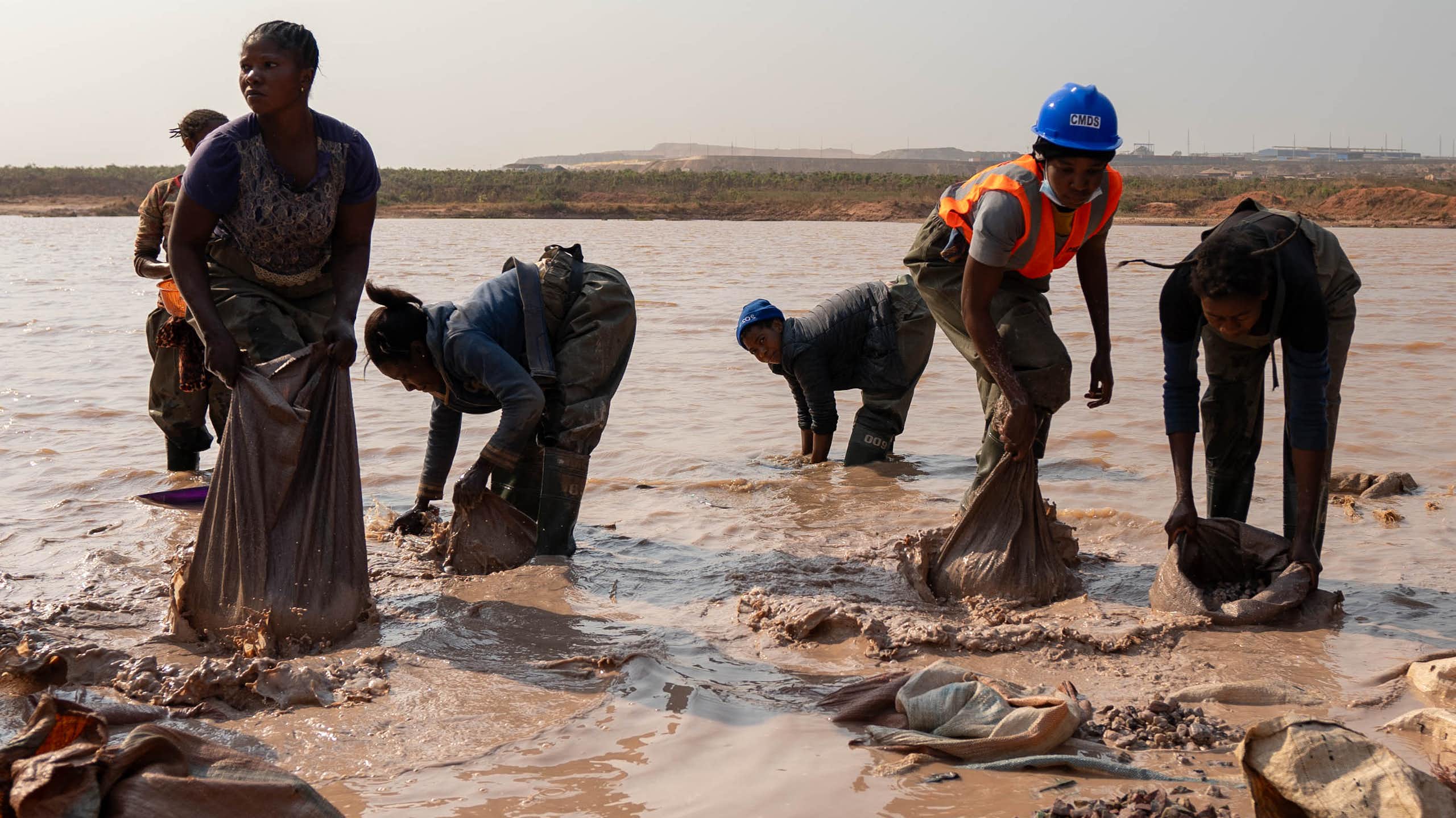 A group of women in a muddy lake, washing something in bags and buckets