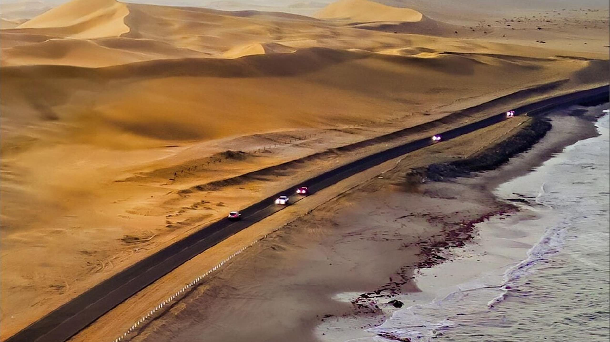 A coastline with a road containing cars and vast sand dunes on the other side of it.