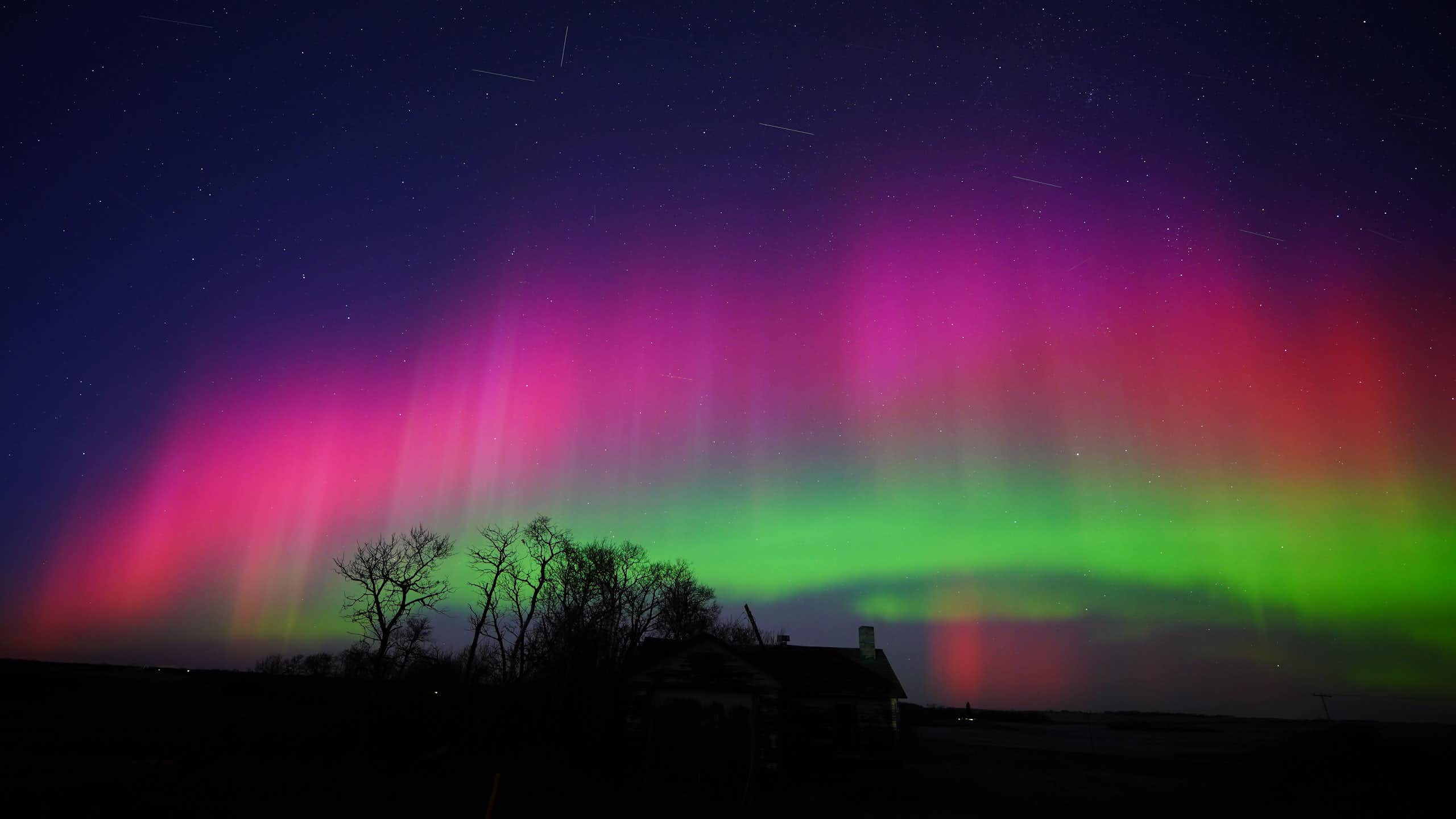 A Starlink train passing through bright green and pink auroras over rural Saskatchewan