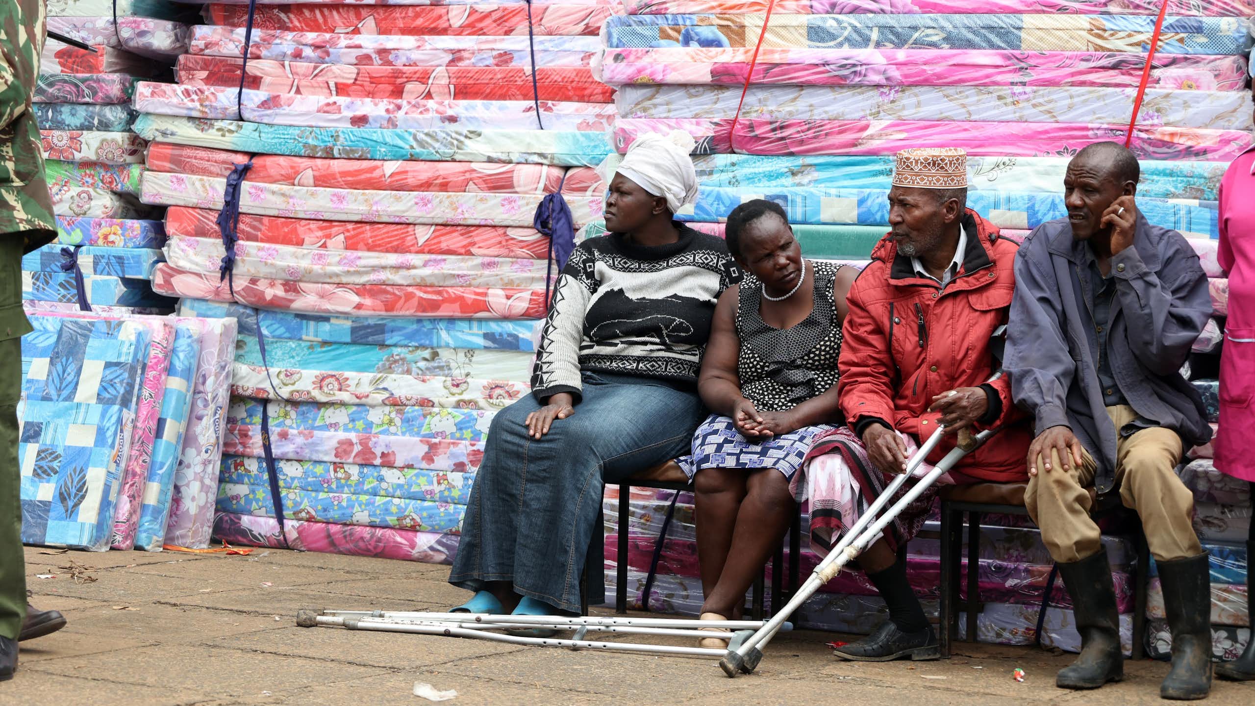 Four people, one of them with crutches, seated next to a pile of mattresses wrapped in plastic