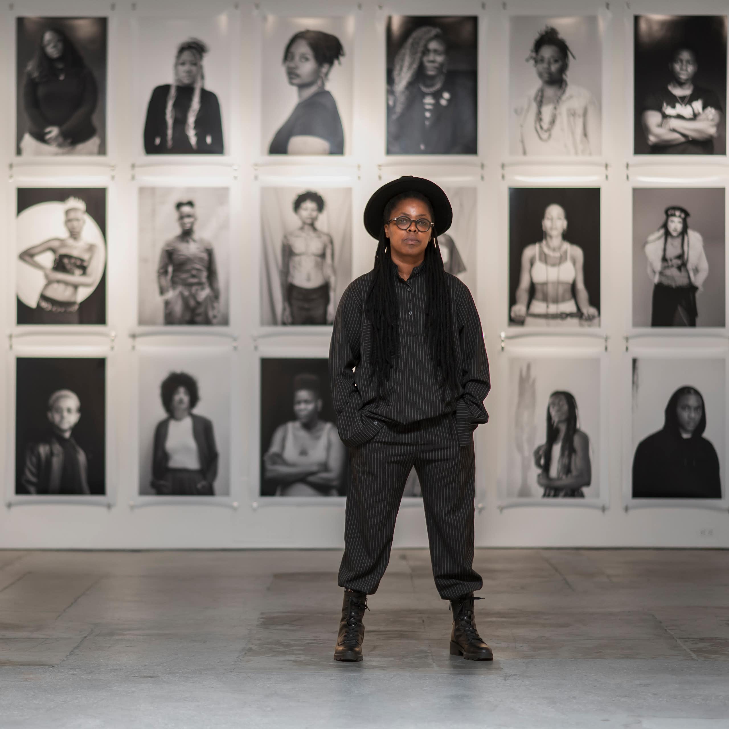 An African woman poses for a portrait against a wall of portraits in black and white.