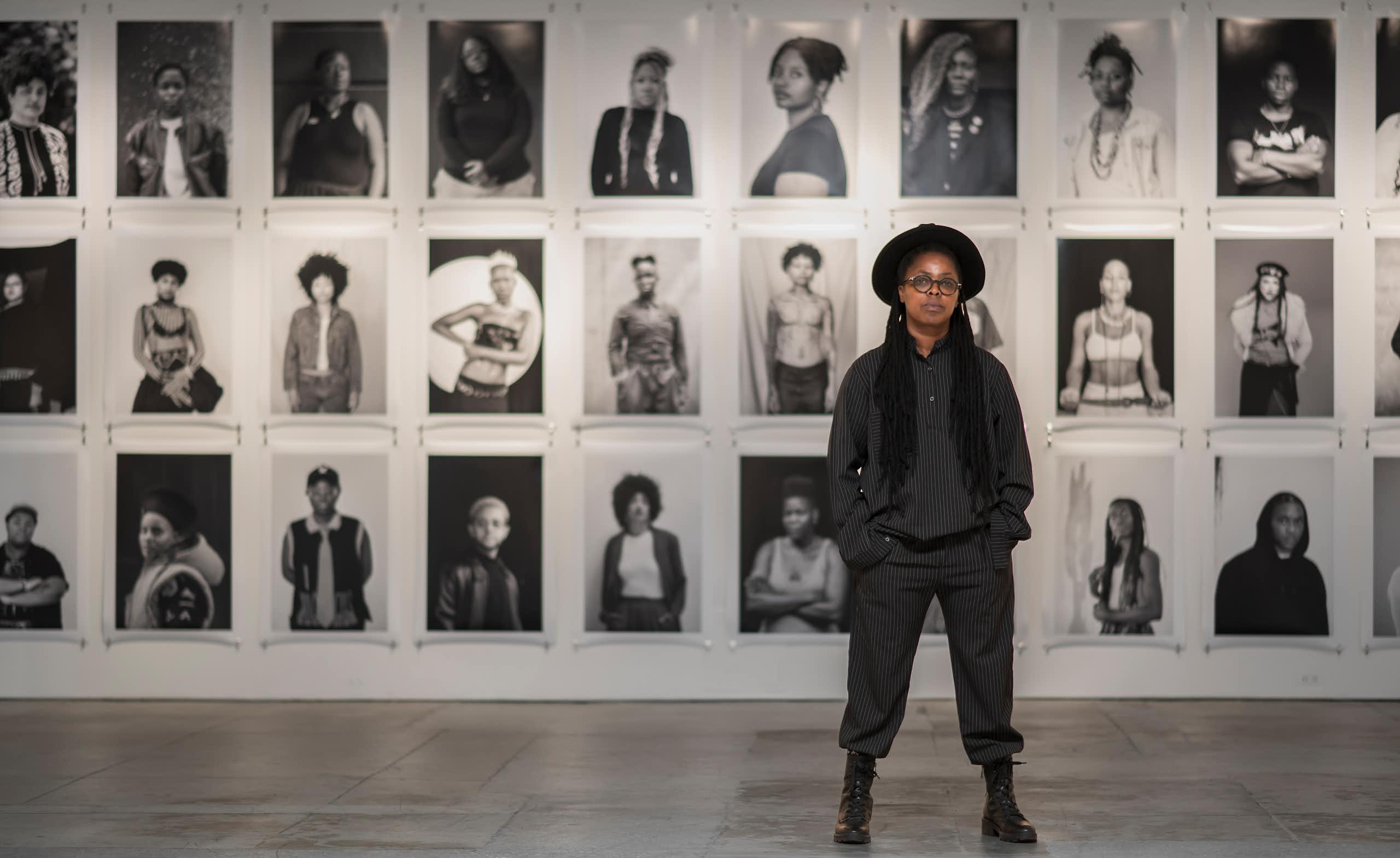 An African woman poses for a portrait against a wall of portraits in black and white.