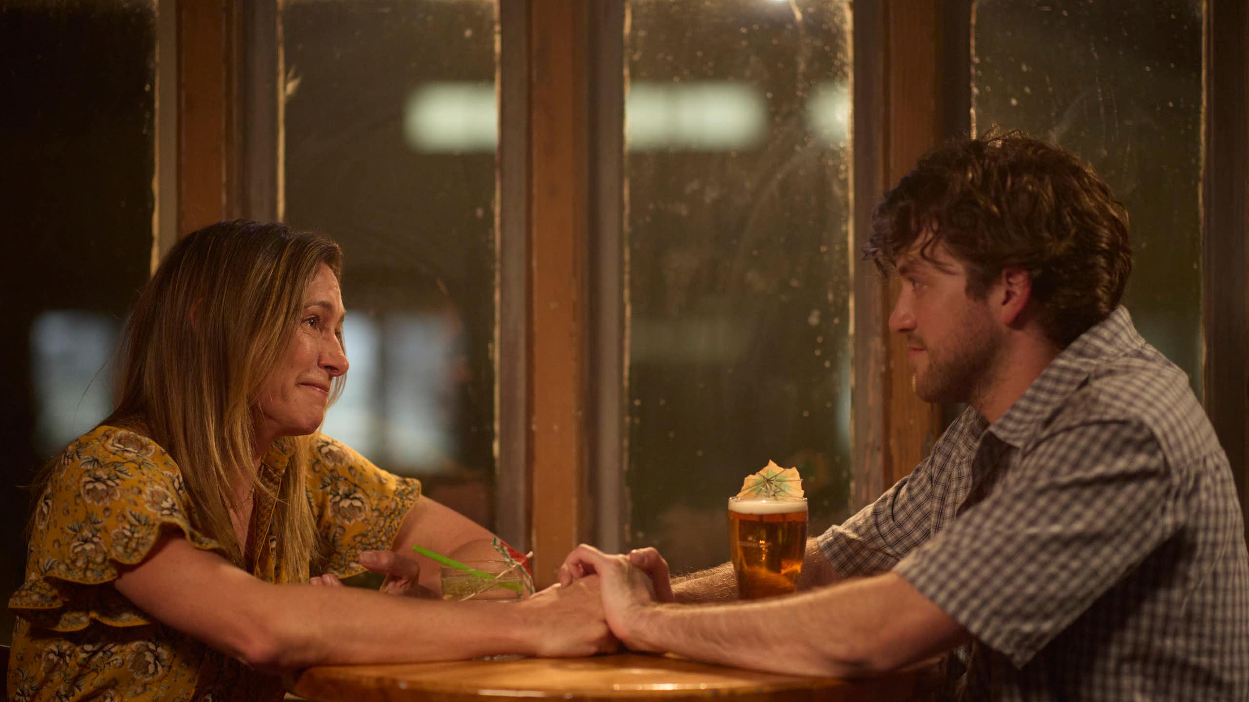 Production image: mother and son hold hands across a table. 