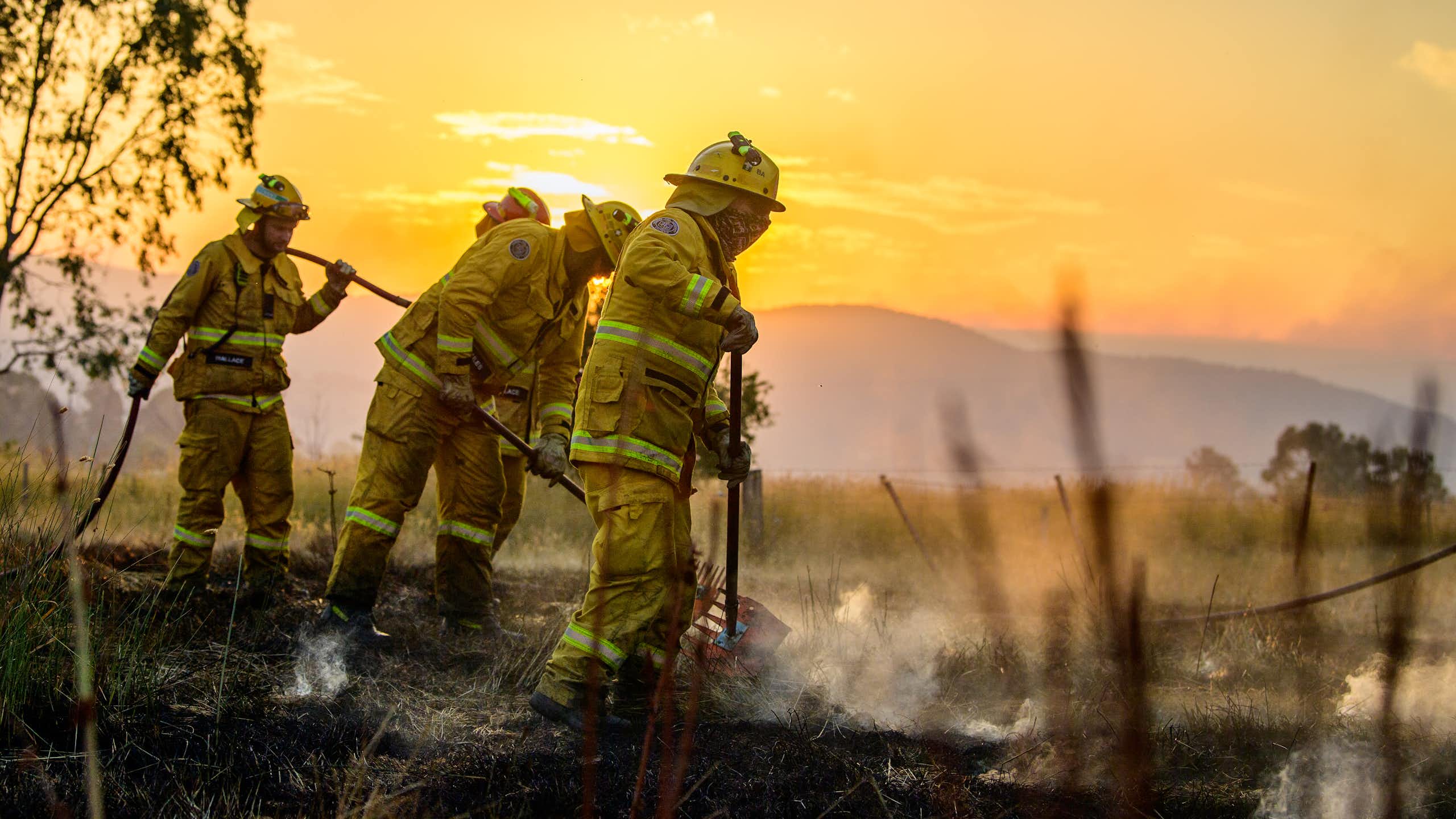 Three firefighters put out a bushfire, with an orange sky in the background.
