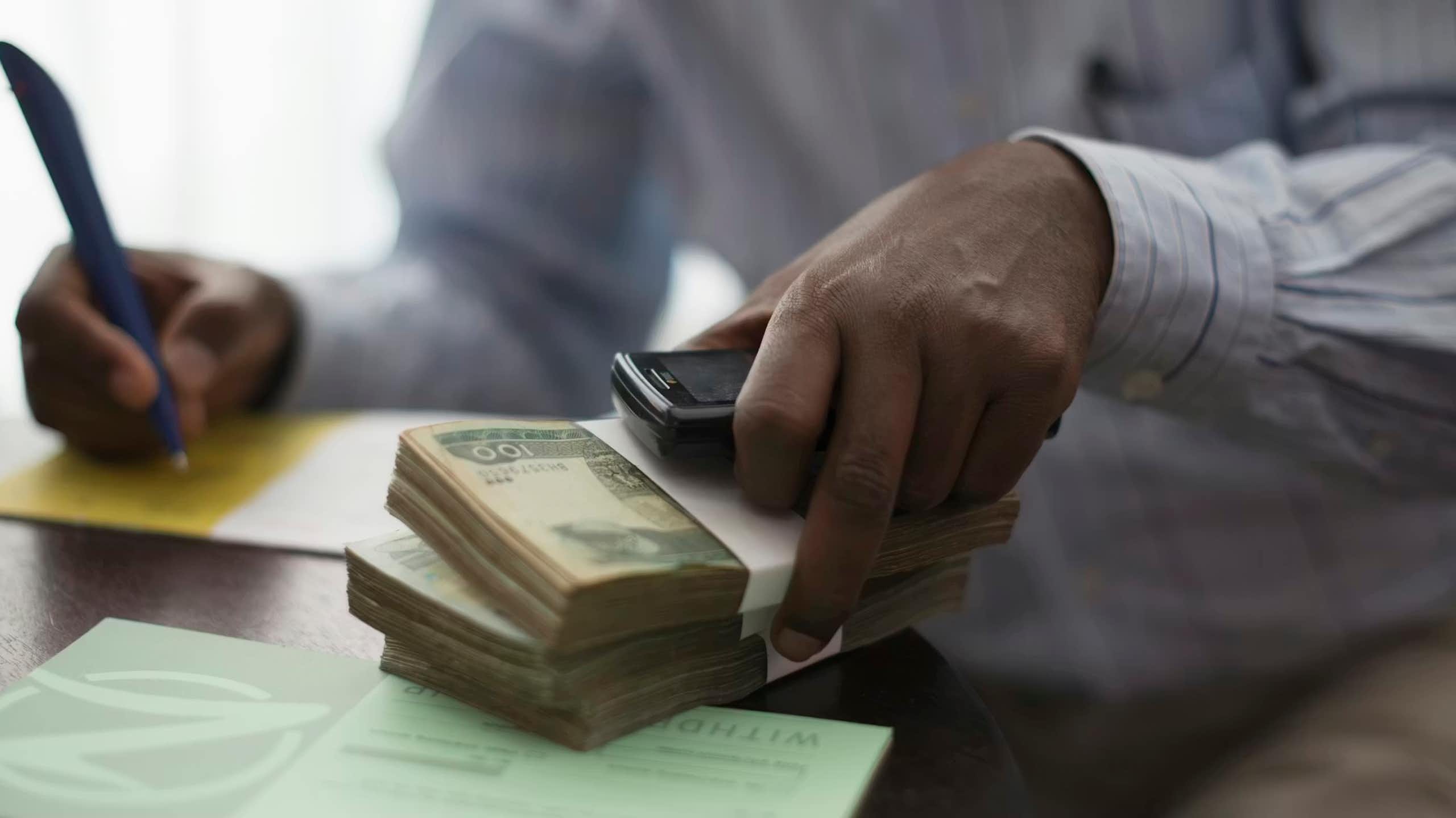 An African man's hands working at a desk, with two piles of cash in one hand he writes on a notepad with the other.