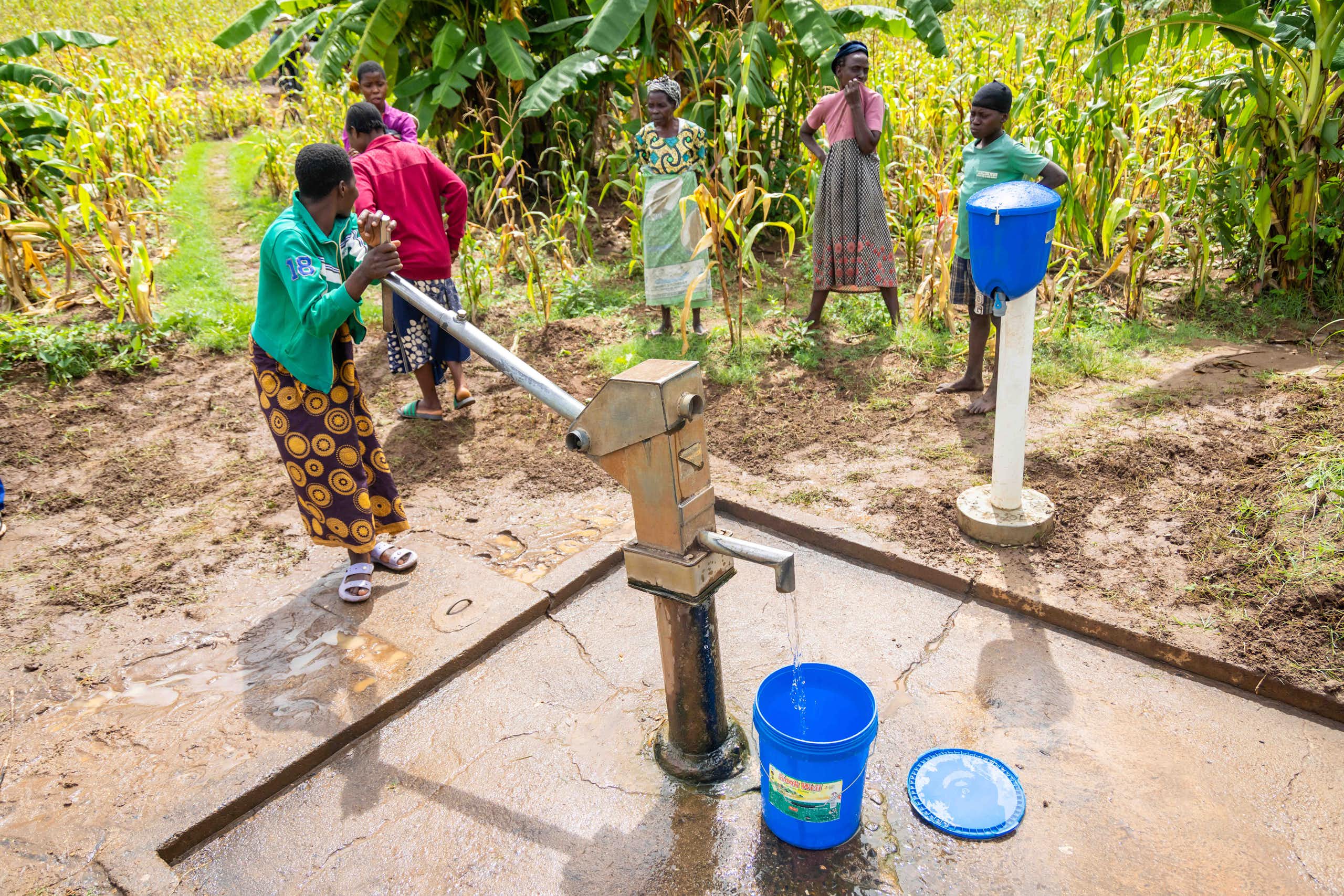 Five people stand with buckets near a tall water pump that is operated by pumping it by hand.
