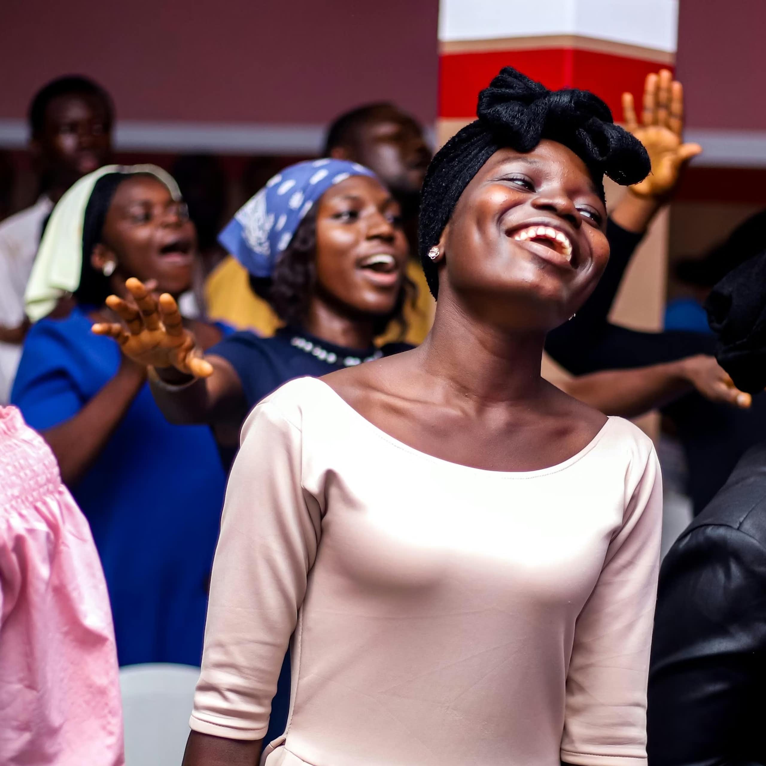 Two young African women smile as they sing, surrounded by other people in a gathering, some with raised arms.