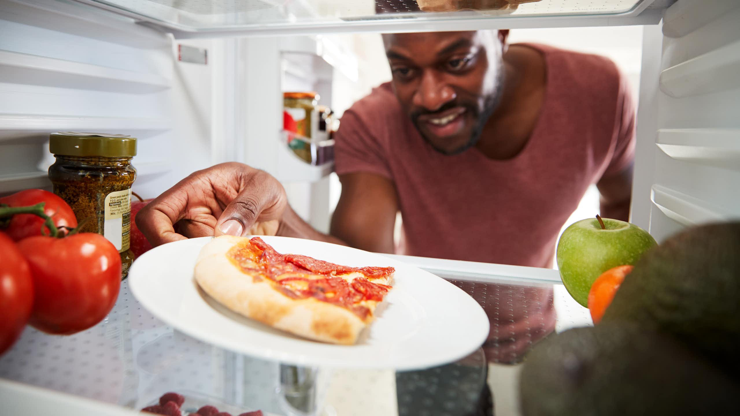 A man reaches into a fridge and grabs a slice of leftover, cold pizza that's sitting on a white plate.