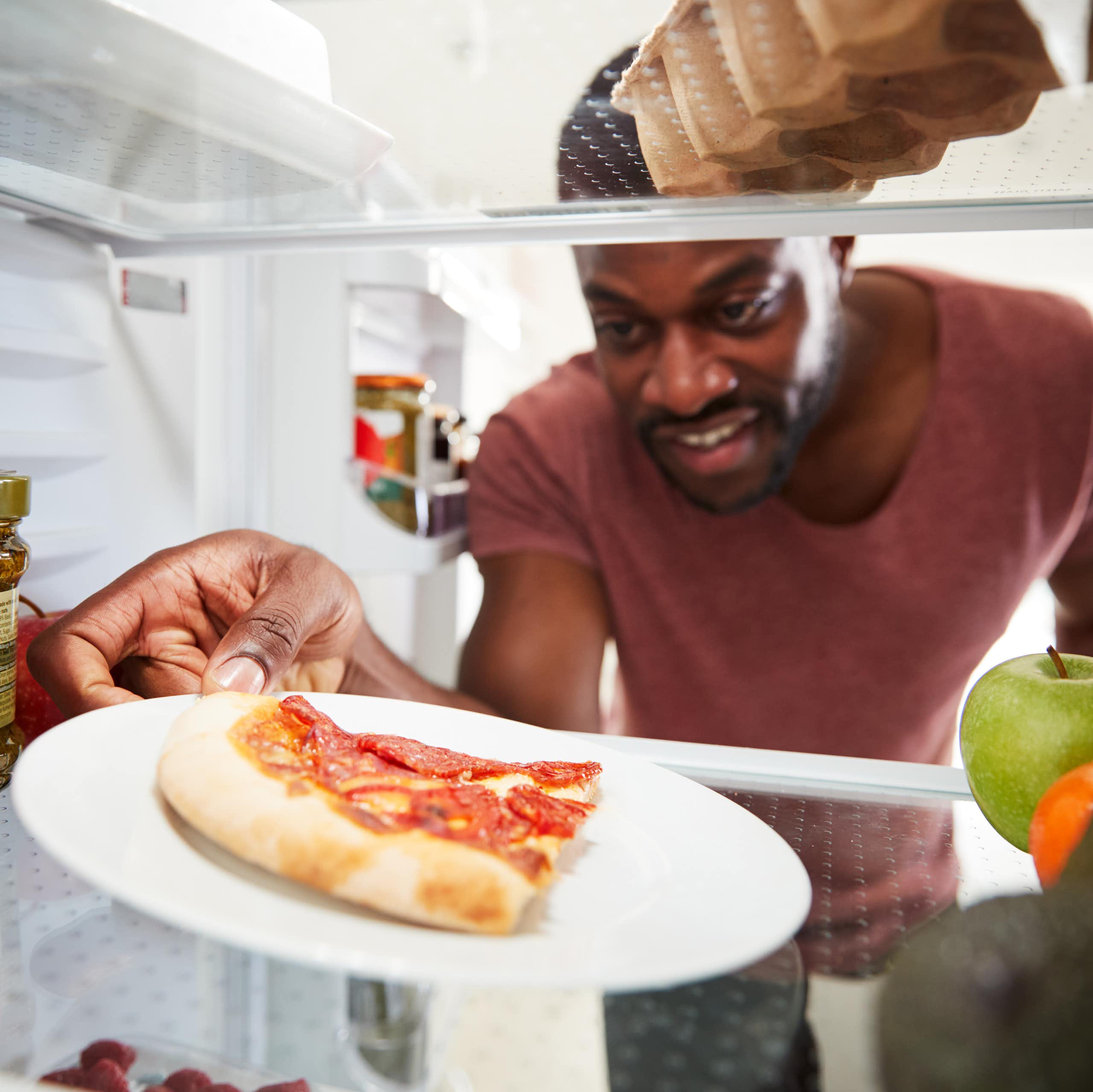 A man reaches into a fridge and grabs a slice of leftover, cold pizza that's sitting on a white plate.
