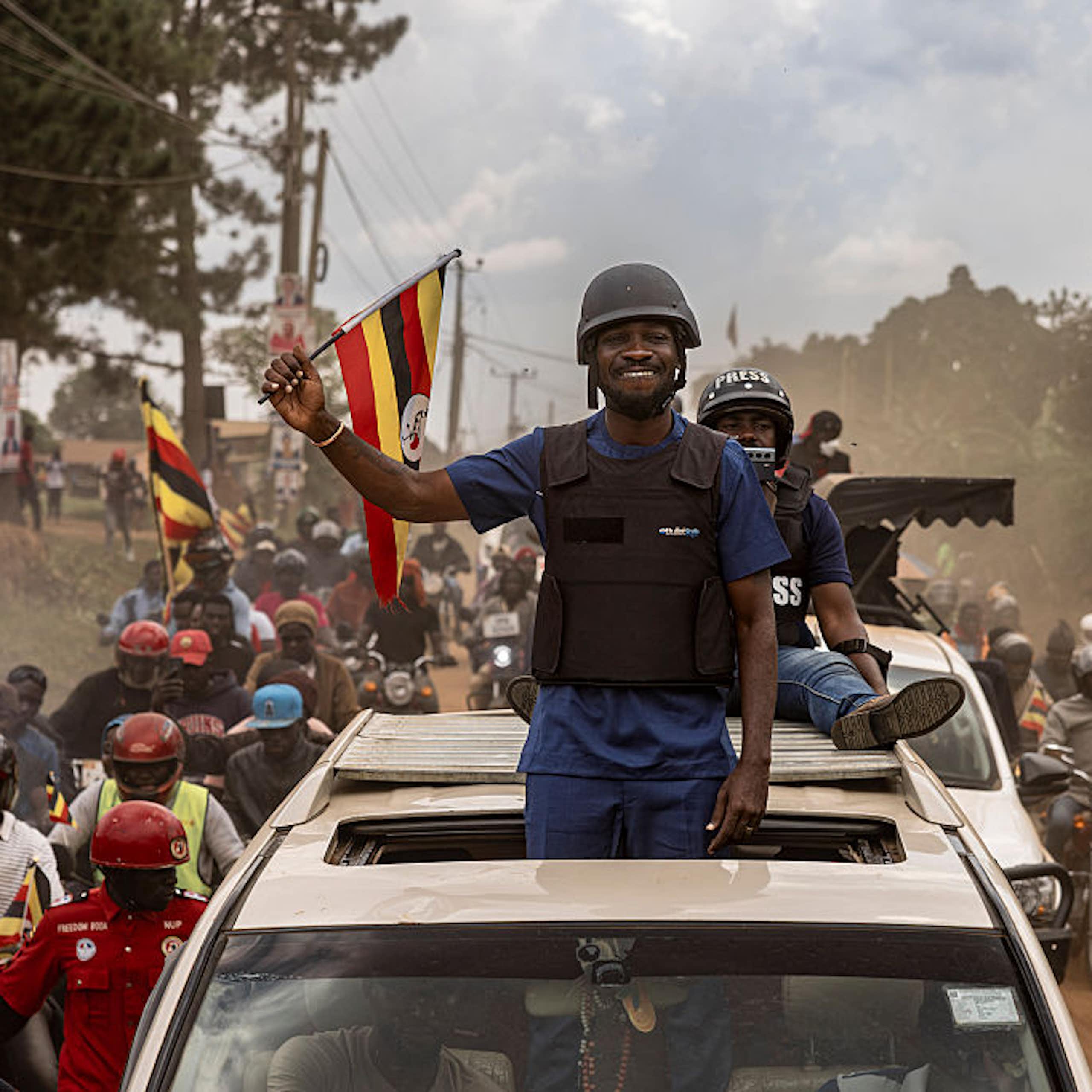 A man wearing a bulletproof vest standing from the sunroof of a vehicle and waving to a crowd of people, some of whom are waving flags.
