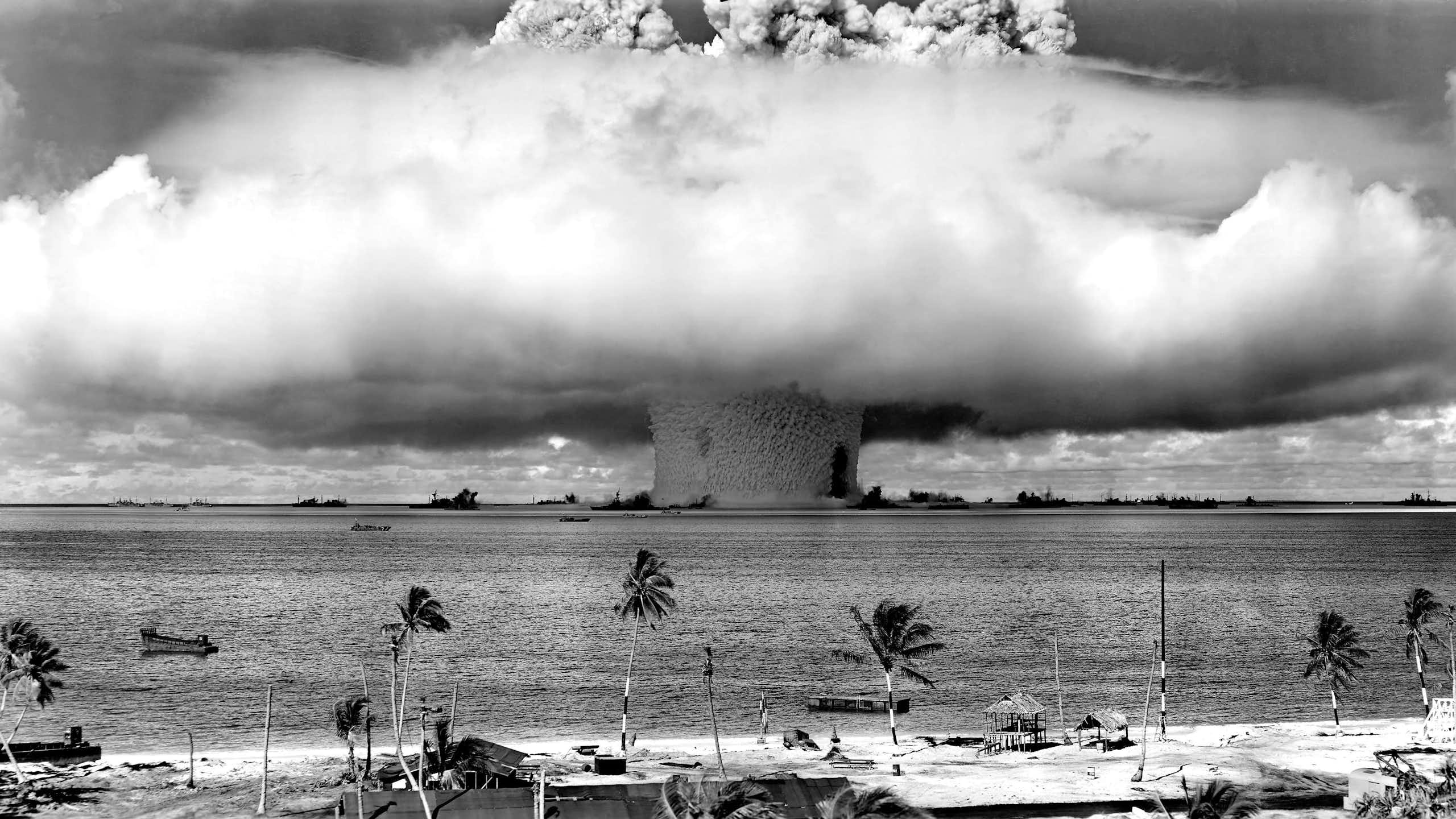 A large mushroom cloud spreads above a beach and water.