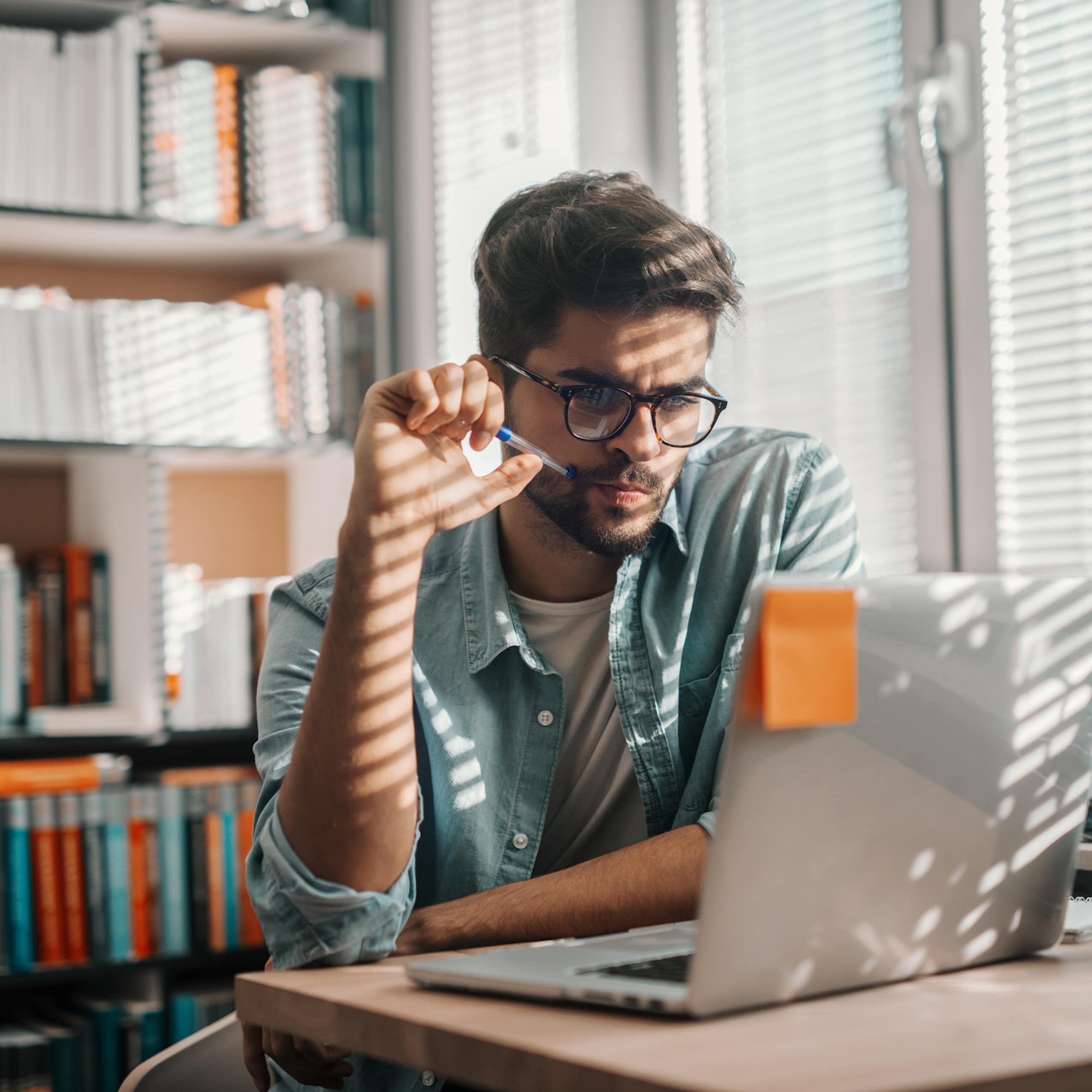 A young man looks at a computer screen. There is a bookshelf behind him. Filtered light comes through blinds to his side. 