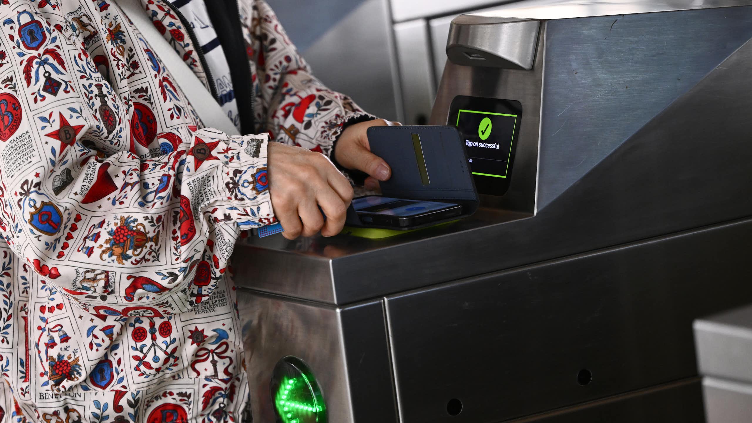 A woman's hands holds a phone over a ticket scanner at a train station turnstile.