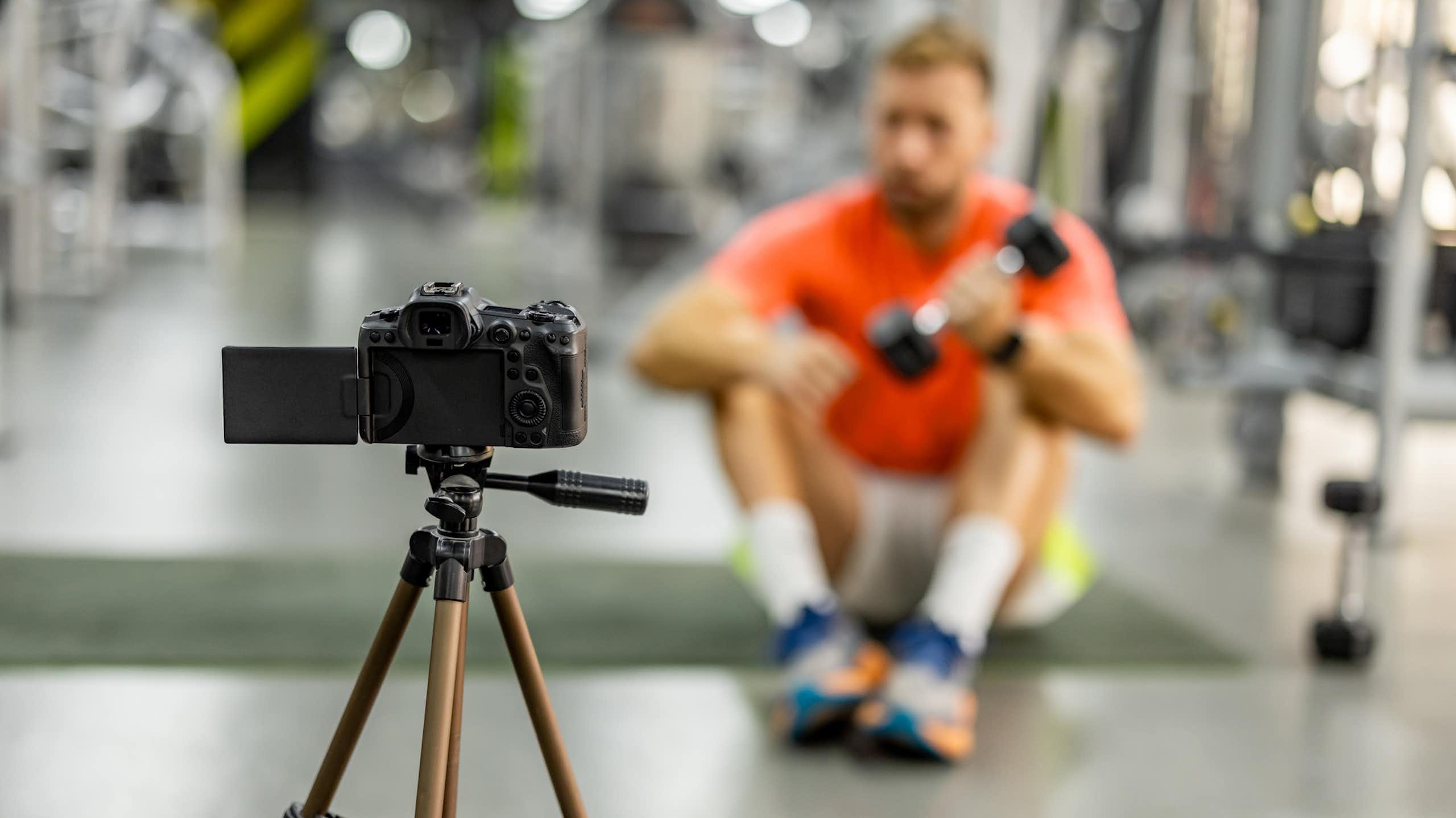 A blurry image of a man in an orange shirt lifting a weight as he sits on the floor in front of a camera tripod.