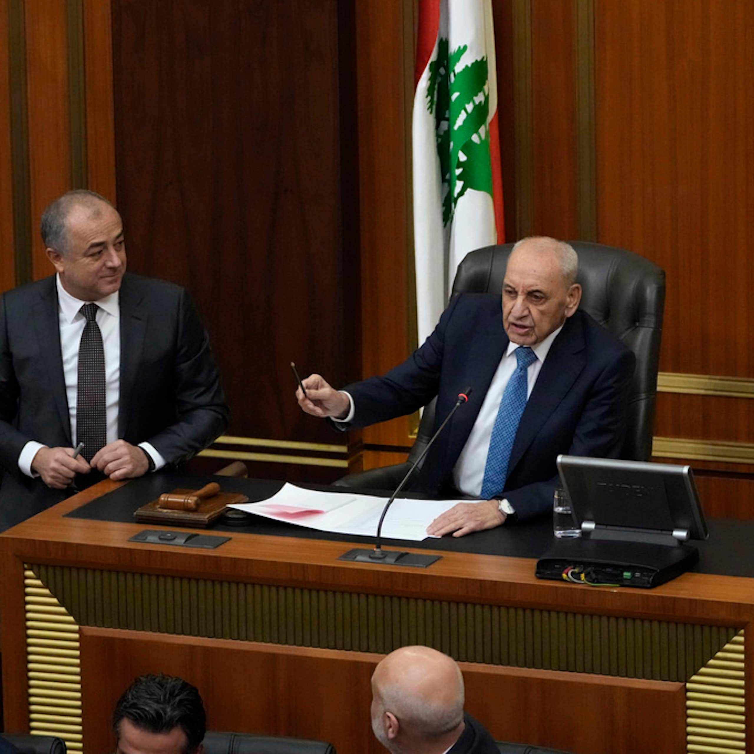 A men in a suit gestures from his speaker's chair.