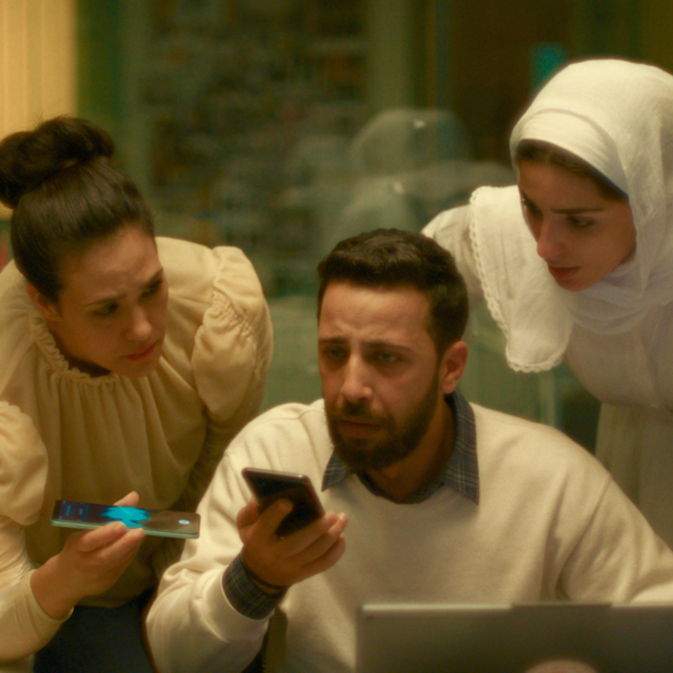 Three women, one in a headscarf, stand anxiously above a man seated with a phone in his hand.